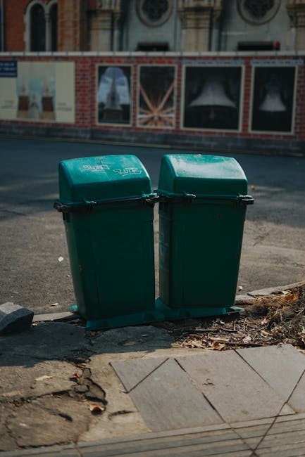 Two green plastic wheelie bins with black handles and locking lids are positioned side by side on a paved sidewalk curb, adjacent to a rough patch of ground with scattered leaves and debris. The bins are made of durable, textured plastic and appear to be used for general waste collection, with some white markings or labels visible on the lids. They are placed in an urban environment, with a street behind them showing a paved surface and a blurred background of a building featuring a red brick facade, large display windows, and decorative architectural details. The scene is illuminated by natural daylight, casting soft shadows on the ground and reflecting slightly on the plastic surfaces of the bins. The setting suggests a typical roadside location for private rubbish disposal, aligning with services provided by companies like Rubbish Collection Barnes for on-site waste removal and alternative disposal options outside local authority collection areas.