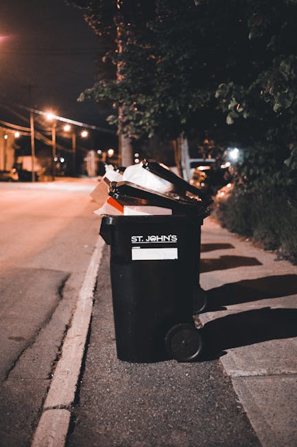 A black wheeled rubbish bin with a rectangular white label and the label 'St. JOHN'S' printed on it, positioned on a sidewalk beside a street at night. The bin is overflowing with various discarded cardboard boxes, paper, and plastic waste, some stacked or leaning out over the rim. The surface of the bin shows signs of use, with a slightly textured finish. Behind the bin, a dark hedge runs parallel to the pavement, and a large tree with leafy branches extends overhead, casting shadows. In the background, streetlights illuminate the empty street, highlighting utility poles and distant parked cars. The scene suggests an informal or private waste collection effort, characteristic of independent rubbish removal services, with a focus on domestic or small-scale clean-up work. The overall lighting is warm and subdued, emphasizing the nighttime environment and the practicality of waste disposal in a residential area, as managed by local rubbish collection services such as Rubbish Collection Barnes.