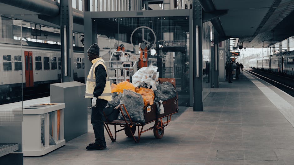 A person wearing an orange protective suit and a white glove is holding a blue plastic rubbish bag by its twisted top, which is partially gathered and tied. The bag appears to be filled with waste, giving it a slightly bulging shape. The individual's hand grasps the bag firmly, suggesting they are in the process of collecting or transporting waste materials. The background is blurred, indicating an outdoor environment, possibly near a waste collection site or a rubbish disposal area. This scene aligns with professional rubbish removal activities, highlighting safe handling and waste containment. The blue rubbish bag's material is thin and glossy, reflecting light slightly, with folds and creases visible along its surface. The orange suit and white glove contrast with the blue bag, emphasizing the controlled handling of waste, a common practice in private rubbish collection services like those offered by Rubbish Collection Barnes. The overall setting underscores the importance of proper waste management and hygienic waste handling in a professional context, supporting the theme of alternative rubbish disposal methods without specifying the exact environment.