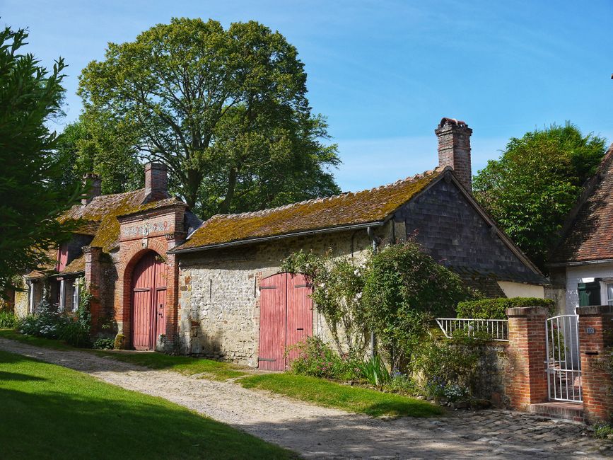 A rustic stone building with a weathered, moss-covered tiled roof, situated in a green rural setting. The structure features pink-painted wooden doors and shutters, with some doors leaning slightly, suggesting recent or ongoing clearance work. To the right, a white metal gate leads to a small garden area with low brick walls and lush shrubbery. In the background, a tall leafy tree rises above the building, casting dappled shade over the scene. The gravel pathway in the foreground runs alongside the building, bordered by well-maintained grass and a neatly kept garden. The environment appears peaceful and quiet, indicative of a private property undergoing interior or exterior clearance, possibly managed by a local rubbish removal service such as Rubbish Collection Barnes, which specializes in discreet and efficient waste disposal for rural or domestic settings. Natural lighting highlights the textures of the aged stonework, moss, and weathered wood, emphasizing the authentic, well-preserved character of the countryside building, aligning with the context of alternative waste handling solutions outside conventional municipal rubbish collection.