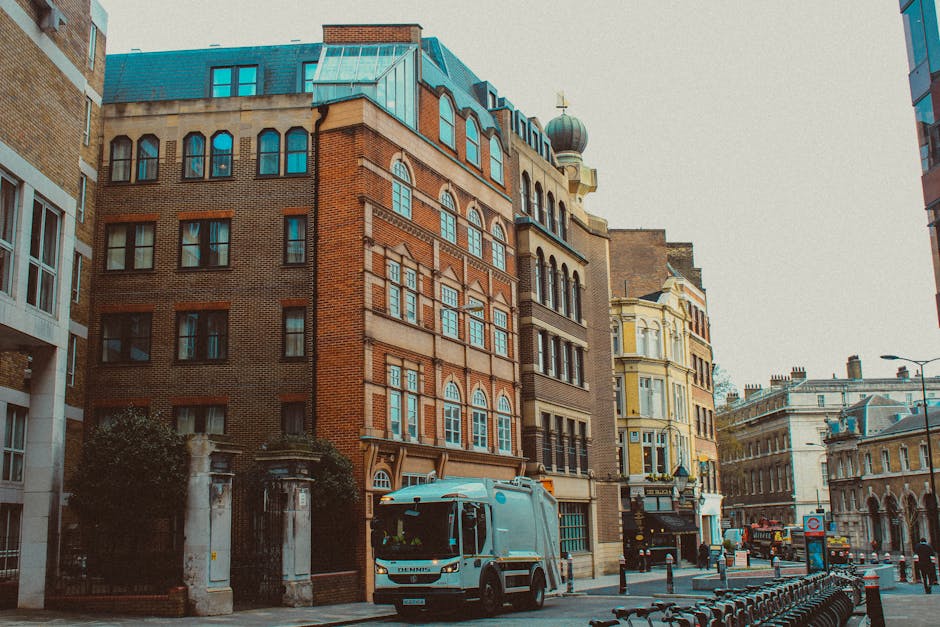 A white refuse collection vehicle parked on a city street in front of a row of historic brick and stone buildings with varied architectural styles, including arched windows, decorative facades, and a domed roof. The vehicle is positioned near the curb, with its rear end facing the viewer, and is likely used by Rubbish Collection Barnes for waste collection or disposal. The street is lined with metal bollards and a bicycle rack, and the scene is illuminated by natural daylight with an overcast sky overhead, creating a neutral, professional atmosphere typical of urban rubbish removal services in a central location. The surroundings include a mix of older and modern buildings, reflecting the area's architectural diversity and hinting at alternative waste handling options for private collection or on-site clearance, as carried out by trusted waste management providers.
