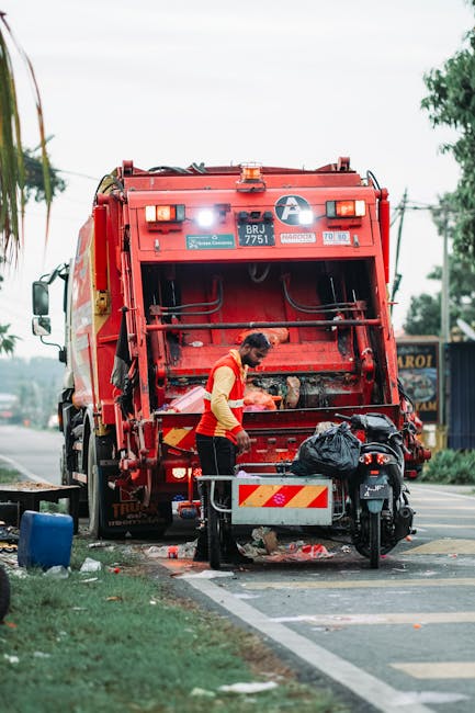 A sanitation worker wearing a yellow high-visibility vest and dark trousers operates a large red rubbish collection vehicle, which is positioned on the side of a street or road. The rear of the vehicle features an open hopper or compaction mechanism, with visible hydraulic components and a metallic finish, designed for waste compaction and disposal. The worker appears to be managing waste or equipment, with a black plastic bag placed nearby on a small metal platform attached to the vehicle. The environment includes a pavement or curb, with some loose rubbish scattered on the ground below. In the background, there are trees and utility poles along the roadside, with some distant signage and possibly a shop or commercial building. The lighting indicates overcast or diffused daylight, providing neutral, natural illumination to the scene. This image aligns with context of private waste collection or on-site rubbish removal services, as provided by companies such as Rubbish Collection Barnes, reflecting independent waste management methods rather than municipal collection systems.