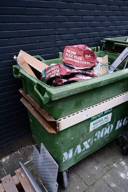 A large green plastic rubbish bin filled with several red and black bags of garden waste or discarded materials, placed outdoors near a dark brick wall. The bin is situated on a paved area and is positioned on top of a white disposal or storage container, which is partially visible beneath it. The bags inside the bin are branded with text indicating they contain garden or waste materials, and the filling appears to be full, with some bags slightly leaning or stacked. The environment suggests a residential or commercial property, where independent waste collection might be utilized as an alternative to public council services. Visible in the background are other waste containers and some wooden blocks on the ground, with natural outdoor lighting illuminating the scene. This setup is typical for private rubbish disposal or on-site clearance, provided by waste management specialists like Rubbish Collection Barnes, supporting effective rubbish removal in the area.